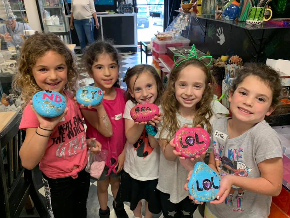 Children happily painting clay with pink and blue colors in a studio. They are adding fun words to their creations, enjoying the creative activities